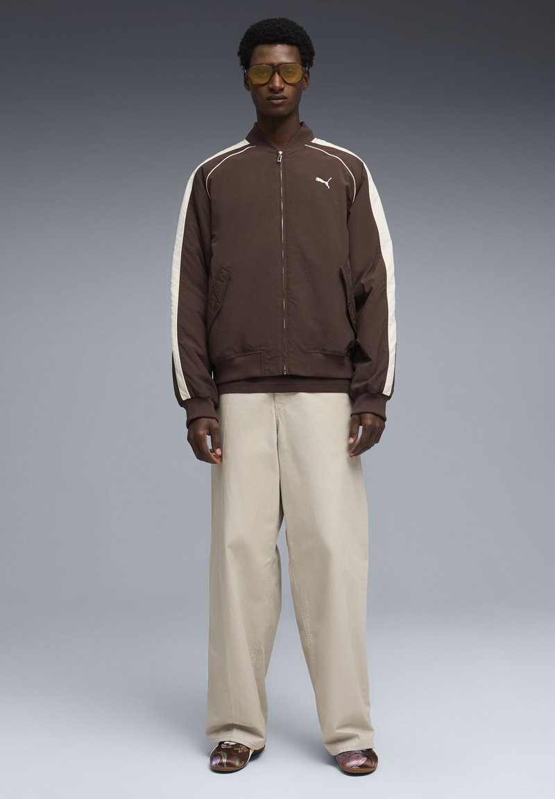 Νεαρός άνδρας φοράει καφέ αθλητικό μπουφάν Puma με λευκές ρίγες, μπεζ παντελόνια με φαρδύ πόδι, γυαλιά με κίτρινο φίλτρο και παπούτσια με σχέδιο σε γκρι φόντο.