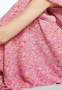 Bras nu reposant sur un tissu rose avec de petits motifs floraux blancs et orange, montrant la texture et la légèreté du matériau.