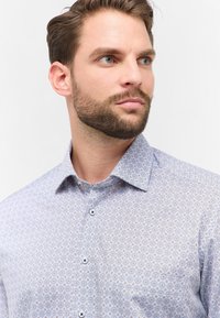 Camicia azzurra chiara con bottoni, caratterizzata da un motivo geometrico in blu scuro e arancione, in tessuto di cotone, colletto aperto e bottoni a contrasto.