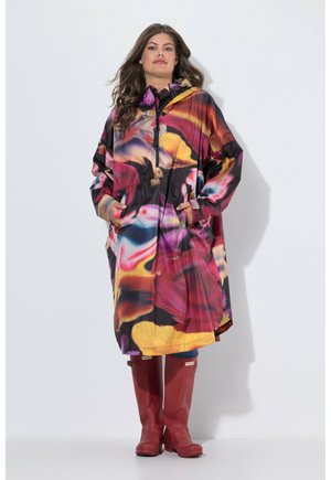 Femme portant un imperméable coloré à motifs, les mains dans les poches et des bottes rouges montant jusqu’aux genoux, debout sur un fond blanc uni.