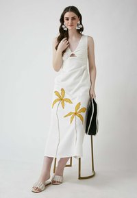 Weißes Leinenkleid mit gelber Palmenstickerei, Ausschnittdetail an der Brust und lockerer Passform. Getragen mit beigen Rüschensandalen.