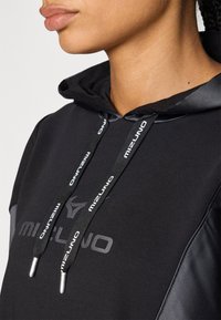 Svart hettegenser med snor med sølvfarger, strukturert stoff og en subtil logo på brystet. Glatte detaljer på skuldrene.