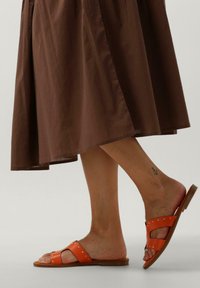 Oranje slide sandalen met twee brede banden, voorzien van zilverkleurige studs en een gestructureerde afwerking. De zool is plat en gemaakt van een lichtbruine stof.