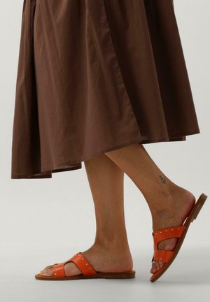 Orangefarbene Rutschsandalen mit zwei breiten Riemen, verziert mit silbernen Nieten und einer strukturierten Oberfläche. Die Sohle ist flach und aus einem hellbraunen Material gefertigt.