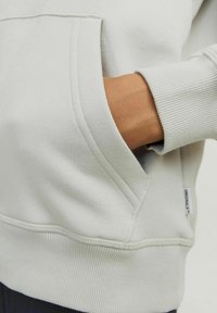 Lichtgrijze sweatshirt met een kangoeroezak, ribgebreide manchetten en zoom. De stof lijkt zacht met een gladde textuur. Label met de tekst "ORIGINALS."