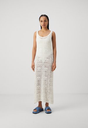 Weißes Häkelkleid mit geometrischen Mustern und design ohne Ärmel; Modell trägt blaue Sandalen mit gekreuzten Riemen.