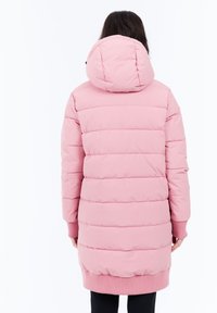 Persona vestida con un abrigo de invierno largo y acolchado de color rosa con capucha, de espaldas frente a un fondo blanco liso.