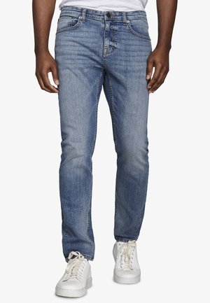 Schmal geschnittene blaue Jeans, getragen von einer stehenden Person, kombiniert mit weißen Turnschuhen und einem weißen Hemd, die Hände locker an den Seiten.