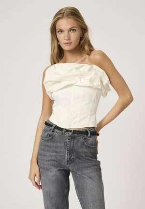 Žena v bielej off-shoulder volánovej blúzke zastrčenej do čiernych vysokých džínsov s tenkým čiernym opaskom.