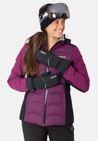 Lila gepolsterte Skijacke mit schwarzen Akzenten, kombiniert mit schwarzen Handschuhen. Model trägt dunkle Skibrille und ein schwarzes Stirnband mit einem Logo.