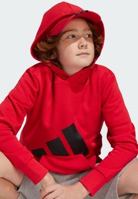 Rotes Kapuzensweatshirt mit einer Vorderang Tasche, das ein großes schwarzes Logo aufweist. Der Stoff scheint weich zu sein. Eine passende rote Mütze vervollständigt den Look.