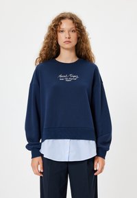 Sudadera color azul marino de corte amplio, con el logotipo bordado "Saint Tropez". Usada sobre una camisa de rayas azul clara, con mangas largas y dobladillo acanalado.