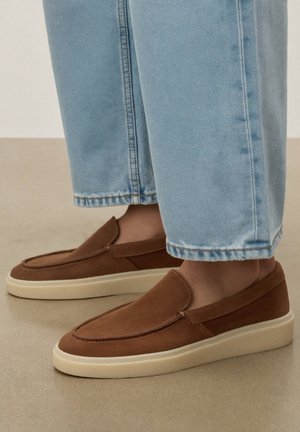 Pantalon en denim bleu clair raccourci au-dessus de chaussures en daim marron à enfiler avec des semelles blanches épaisses sur un sol beige.