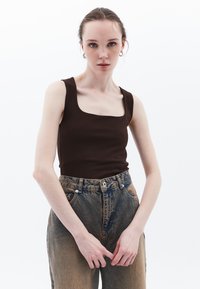 Top aderente color marrone con scollatura quadrata, abbinato a jeans in denim blu sbiadito a vita alta. La modella è in piedi con le mani unite.