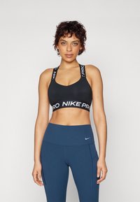 Crna sportska brusnica sa �širokim trakama koja nosi "NIKE PRO" oznaku, uparen sa visokim tamnoplave tajice s bočnim džepovima i malim logotipom.