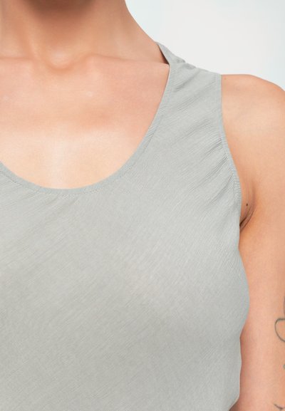 Close-up of a person wearing a light gray sleeveless top, showing neck, upper chest, and partial tattooed arm on white background.