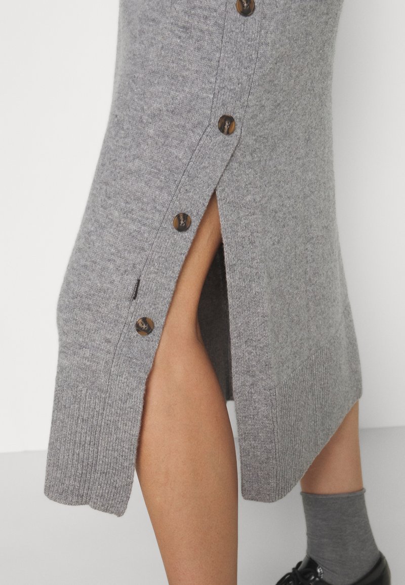 Szara dzianinowa spódnica do kolan z bocznym rozcięciem na guziki, odsłaniającym odkrytą nogę; zestawiona ze szarymi skarpetkami i czarnymi butami na białym tle.