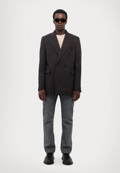 Sort stribet dobbeltbrystet blazer, beige t-shirt underneath, grå jeans, sorte sko og mørke solbriller, stående mod en hvid baggrund.