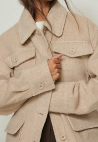 Beige uldjakke med krave, frontknapper og to brystlommer. Blød tekstur med en lidt oversized pasform.