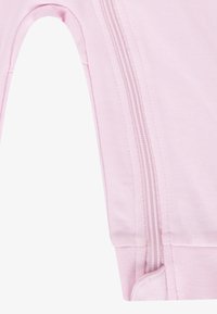 Vêtement rose avec une fermeture éclair sur le côté, doté d'une texture lisse et de poignets élastiques. Le design comprend une zone ouverte pour une meilleure liberté de mouvement.