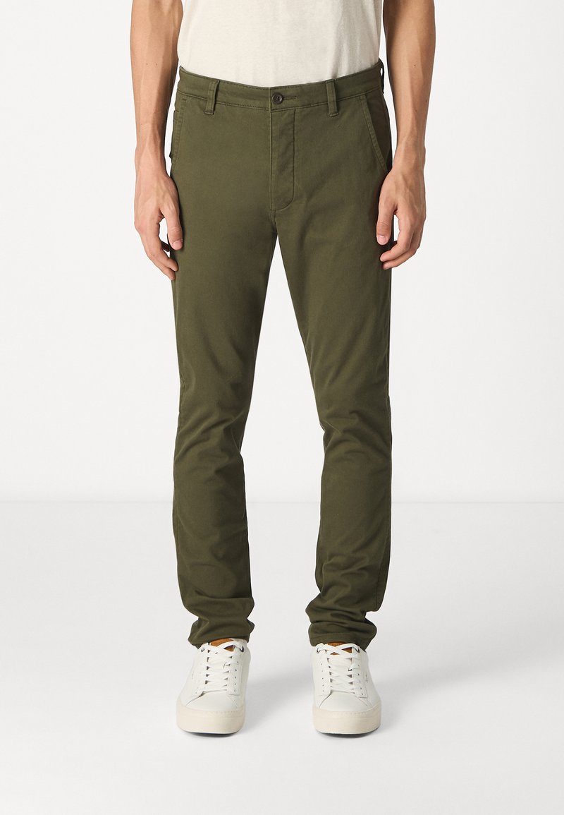 Chinos vert olive en tissu de coton mélangé, avec une coupe slim, des poches latérales et une fermeture à un seul bouton. Associés à des baskets blanches.