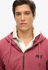 Sudadera con cremallera color burdeos con cordones y bordado del logo. Lleva una camisa blanca con cuello que muestra un forro gris visible. Textura de tela suave.