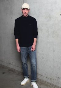 Mann mit weißer Kappe, schwarzem Langarmshirt, blauen Jeans und weißen Turnschuhen, der an einer grauen Betonwand steht.