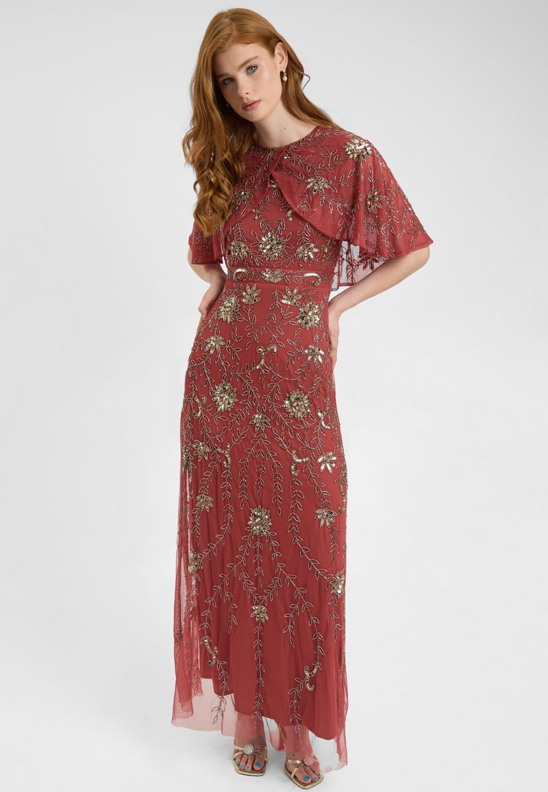 Abito rosso ricamato con fiori e maniche in chiffon. Presenta dettagli intricati in oro, un corpetto aderente e una gonna fluente. Accenti metallici sulle scarpe.