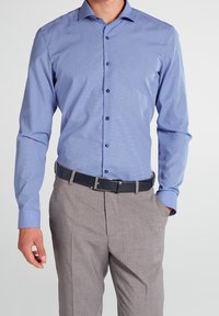 Man wearing a light blue long-sleeve button-up shirt tucked into gray dress pants with a black belt, hand in pocket.