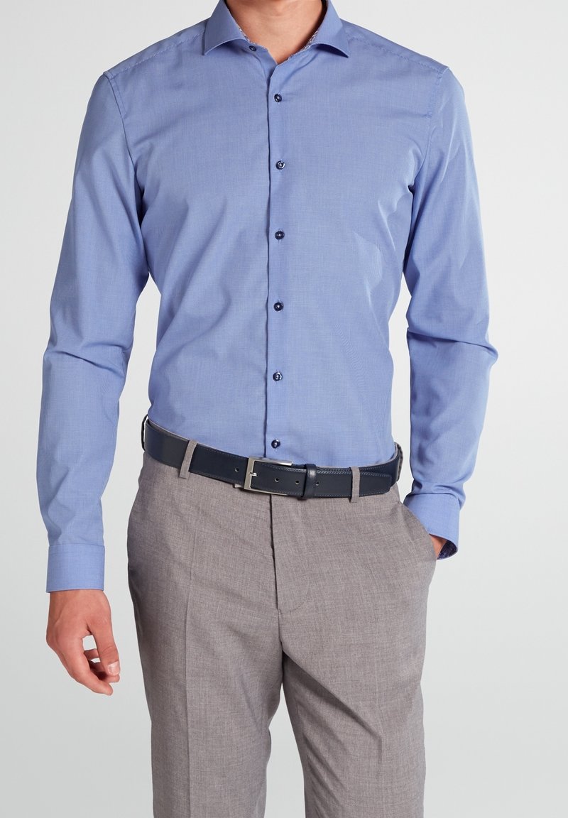 Man wearing a light blue long-sleeve button-up shirt tucked into gray dress pants with a black belt, hand in pocket.