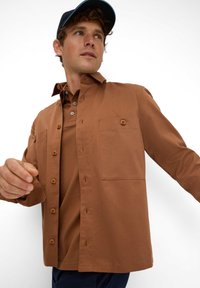 Brun skjorte med knapper og to frontlommer, lavet af bomuldsstof, lange ærmer, moderne pasform og afslappet krave; båret over en brun polo.