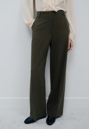 Personne portant un pantalon taille haute à jambes larges vert olive, un chemisier crème à volants et des mocassins noirs, debout avec les mains partiellement dans les poches.