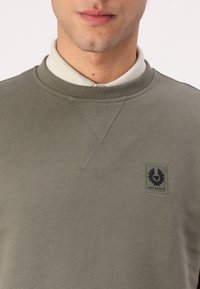 Khaki színű pulóver, kerek nyakkivágással, keresztöltögetéses mintával és sötét emblémával a mellkason, amelyen a "BELSTAFF" felirat található.