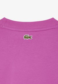 T-shirt in cotone rosa con scollatura a girocollo e un piccolo logo di coccodrillo verde ricamato vicino al collo sul retro.