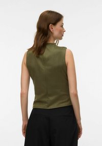 Femme aux cheveux bruns mi-longs portant un haut sans manches vert olive et un pantalon noir, vue de dos sur un fond uni.