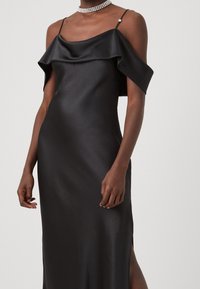 Svart satindress med draperad design som går över axeln, tunna band och slits på sidan. Har en slät textur och en elegant silhuett.