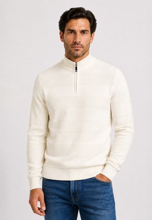 Mand med mørkt hår iført en hvid kvart-zip sweater og blå jeans, stående med den ene hånd i lommen mod en ensfarvet baggrund.