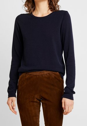 Vrouw draagt een aansluitende marineblauwe trui met lange mouwen en bruine suède broek met elastische tailleband, staand tegen een effen achtergrond.