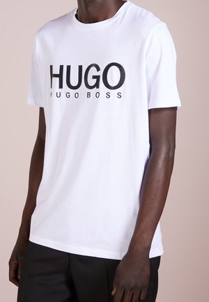 T-shirt en coton blanc avec un grand logo noir "HUGO" et le texte "HUGO BOSS". Manches courtes, col rond et coupe ajustée.