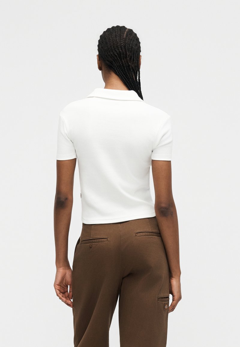 Donna con capelli intrecciati vista da dietro, che indossa una camicia bianca a maniche corte con colletto e pantaloni marroni con tasche.