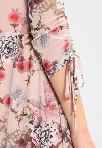 Detalle de un vestido floral con una tela rosa transparente y mangas fruncidas, patrones florales en blanco, rojo y verde, y delicados lazos decorativos.