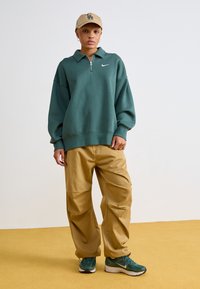 Sweatshirt verde com fecho, logótipo da Nike e corte oversized; combinado com calças largas khaki e sapatilhas verdes. Fundo de chão bege.