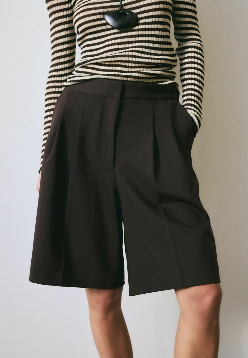 Oseba, ki nosi široke kratke hlače črne barve z visokim pasom do kolen in dolg rokav bež s črnimi črtami ter črno ogrlico z obeskom.