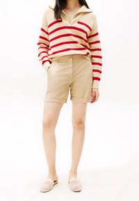 Beige trui met rode strepen, rits hals; beige shorts met omgeslagen zoom; lichte geweven instappers. Eenvoudige, casual outfit.