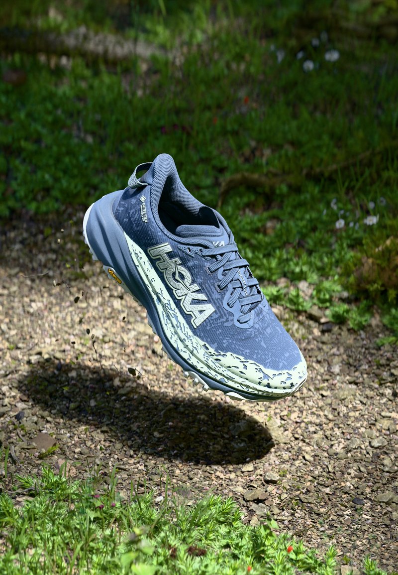 Scarpa da corsa leggera grigia e menta con tomaia testurizzata, logo prominente e suola robusta, progettata per durabilità e supporto su sentieri.