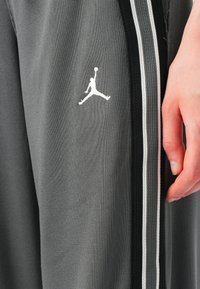 Pelēkas sporta bikses, izgatavotas no teksturēta auduma, ar baltu Jordan logotipu un melnbaltiem sānu rakstiem. Roka daļēji redzama, turot audumu.