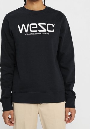 Henkilö, joka pukeutunut mustaan huppariin, jossa valkoinen "WESC"-logo, ja beigen värisiin housuihin, seisoo yksivärisen vaalean taustan edessä.
