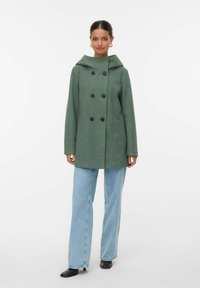 Grøn uldfrakke med dobbeltknaplukning, stor hætte, sidelommer og afslappet pasform, parret med lyseblå jeans og sorte sko.