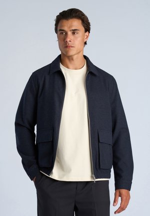 Mand iført mørk navy lynlåstrøje med store forlommer over en lys beige skjorte og sorte bukser, stående mod en bleg grå baggrund.