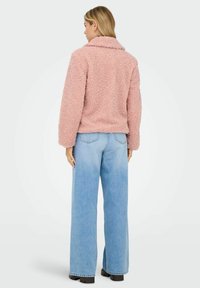 Pink, fluffy, cropped jacket with a soft texture, wide sleeves, and bulky collar, paired with light blue, high-waisted wide-leg jeans.